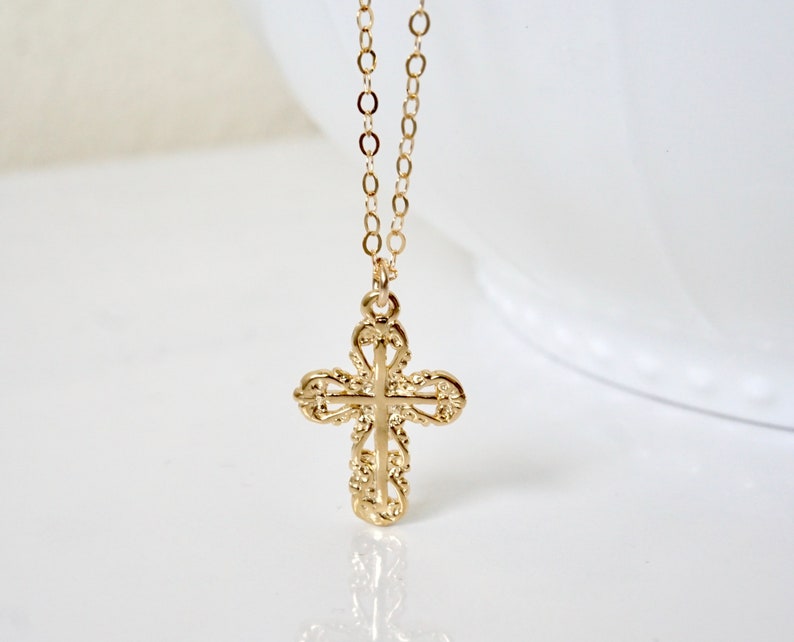 May include: A gold chain necklace with a delicate, ornate cross pendant. The cross is detailed with intricate scrollwork and has a slightly raised center.