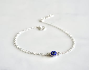 Könnte beinhalten: Ein filigranes Armband aus Silberkette mit einem kleinen, runden Anhänger mit einem blau-weißen Evil-Eye-Design. Das Armband wird auf einer weißen Oberfläche gezeigt, was den Kontrast zwischen der Silberkette und dem leuchtenden Blau des Anhängers hervorhebt.