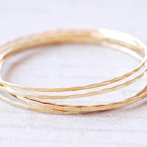 May include: Four gold-colored bangles with a hammered texture. The bracelets are thin and round, and they are stacked together. The bangles are made of a shiny, metallic material.