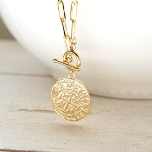 Könnte beinhalten: Goldkette mit einem großen, runden, strukturierten Münzenanhänger. Die Kette hat einen Toggle-Verschluss.