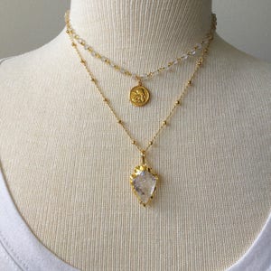Puede incluir: Un collar de oro en capas con un colgante de cristal transparente y un peque&ntilde;o dije de &aacute;ngel de oro.