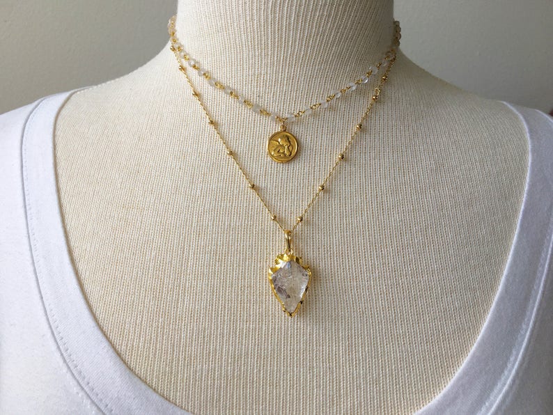 Puede incluir: Un collar de oro en capas con un colgante de cristal transparente y un peque&ntilde;o dije de &aacute;ngel de oro.