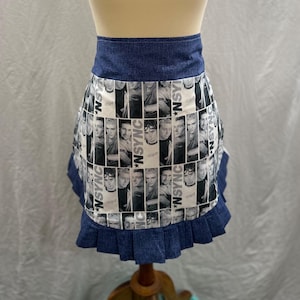 May include: A denim apron with a white and black pattern featuring images of *NSYNC band members. The apron has a denim waistband and a ruffled denim hem. The apron is displayed on a mannequin.