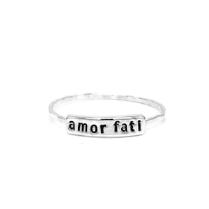 May include: A silver ring with a hammered texture and a rectangular band that says "amor fati" in black lettering.