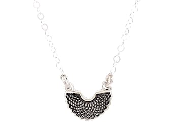 May include: A silver-toned necklace featuring a delicate chain and a small, semi-circular pendant. The pendant has a detailed, dark-colored, intricate web-like pattern etched into its surface, suspended from two small jump rings on the chain.