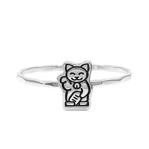 Puede incluir: Anillo de plata con un dije de gato Maneki-neko en blanco y negro. El gato está representado de pie con una pata levantada, un símbolo tradicional de buena suerte y fortuna. La banda del anillo tiene una textura martillada.