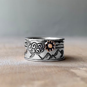 Peut inclure: Une bague tournante en argent avec un motif de montagne et de nuage. La bague est ornée d'une petite fleur dorée.