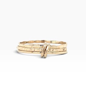 May include: A gold-colored ring featuring a stack of three thin bands with a hammered, textured finish. The bands are joined together by two small, vertical gold-colored wire loops that wrap around the center of the stacked ring structure.