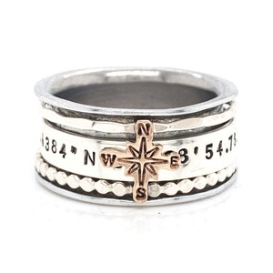 May include: A silver ring with a compass charm and coordinates engraved on the band. The ring is made of multiple bands and has a textured finish.