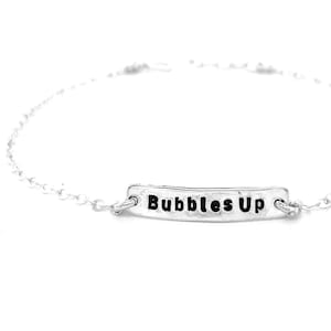 May include: A silver chain bracelet with a silver bar charm that says "Bubbles Up" in black lettering.