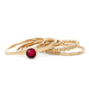 May include: Set of four gold rings, one with a red gemstone, stacked on a white surface.