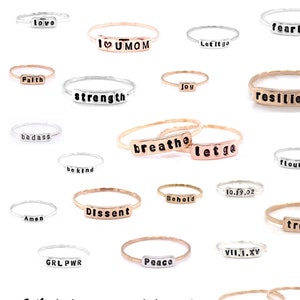 May include: Assortment of silver and gold rings with various words and phrases engraved on them, including "love", "fearless", "I love U MOM", "Let it go", "faith", "joy", "strength", "resilient", "badass", "breathe", "let go", "be kind", "flourish", "10.19.02", "behold", "dissent", "amen", "GRL PWR", "peace", "VII.I.XV", "truth", "Karleigh", and "Claire". The rings are arranged in a random pattern on a white background. The text "What do you want to say?" is written in black cursive at the bottom of the image.