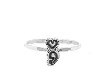 Pode incluir: Um anel de prata com um design de coração e ponto e vírgula. O anel tem uma banda fina e uma superfície texturizada. O coração está posicionado acima do ponto e vírgula, criando uma joia única e simbólica.