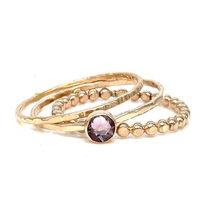 Puede incluir: Un conjunto de tres anillos de oro, uno con una pequeña piedra preciosa púrpura. Los anillos están apilados uno encima del otro.