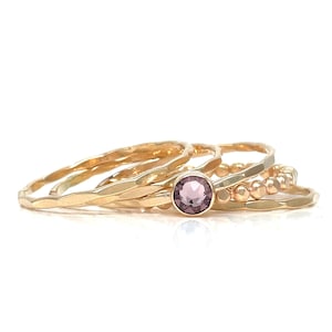 May include: Set of four gold rings, one with a small purple gemstone. The rings are stacked on top of each other.
