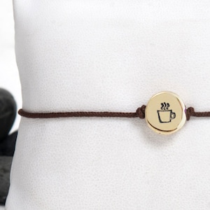 Könnte beinhalten: Ein braunes Kordelarmband mit einem goldenen Anhänger, der eine Kaffeetasse zeigt. Das Armband wird auf einem weißen Kissen präsentiert.
