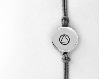 May include: A silver bracelet with a circular pendant displaying a triangle within a circle symbol. The bracelet is strung on a grey cord and is presented on a white cushion. Dark stones are in the foreground.