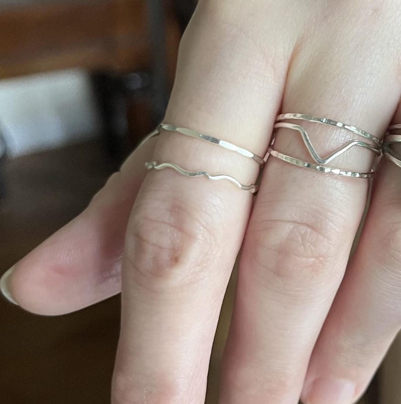 May include: A close-up of a hand wearing four silver rings. The rings are stacked on the finger and are all different styles. The rings are simple and delicate.