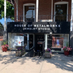 May include: A storefront with a black awning that reads "House of Metalworks Jewelry Studio". The storefront has two large windows with displays of jewelry inside. There are plants in pots outside the store.