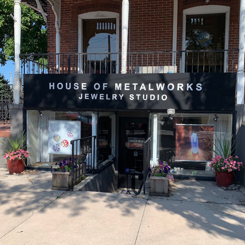May include: A storefront with a black awning that reads "House of Metalworks Jewelry Studio". The storefront has two large windows with displays of jewelry inside. There are plants in pots outside the store.