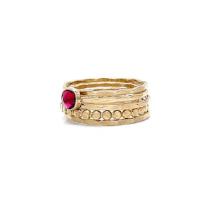 May include: A set of five gold rings with a single red gemstone ring in the centre. The rings are stacked on top of each other.