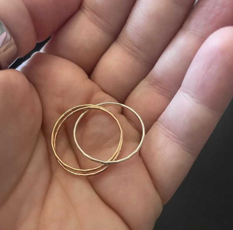 K&ouml;nnte beinhalten: Nahaufnahme von drei d&uuml;nnen Ringen: zwei goldfarben und einer silberfarben. Die Ringe sind &uuml;bereinander gestapelt, wodurch ein geschichteter Effekt entsteht. Die goldenen Ringe haben eine strukturierte Oberfl&auml;che, w&auml;hrend der silberne Ring glatt erscheint. Die Ringe werden in einer Hand gehalten.