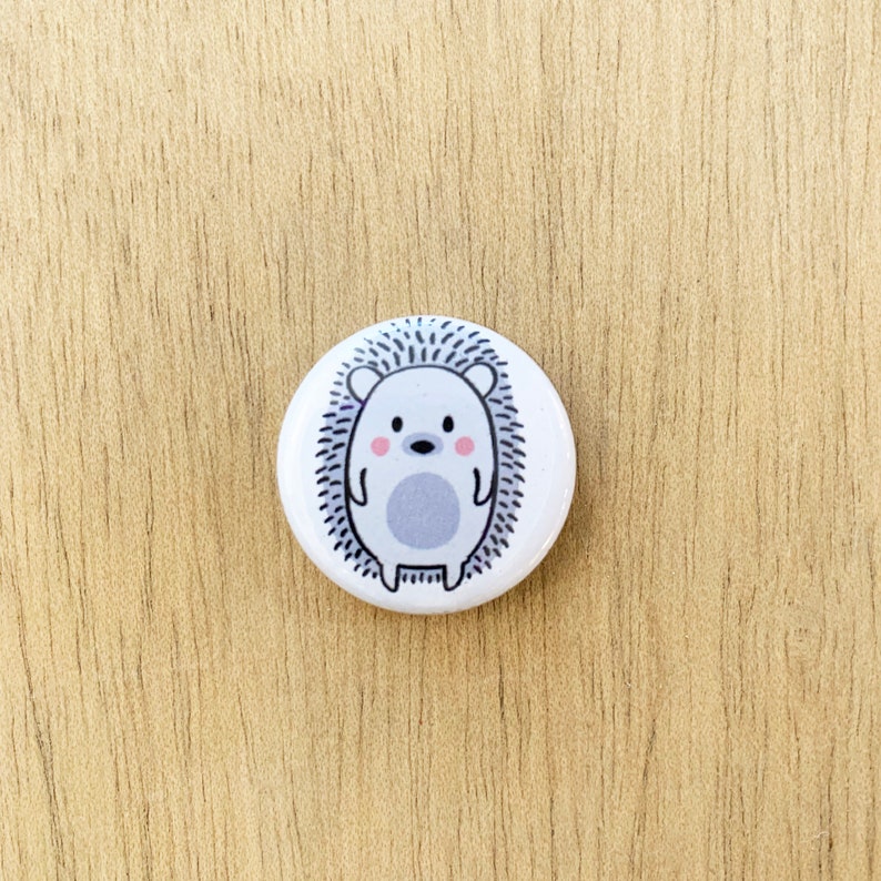 May include: A white button with a cartoon hedgehog design. The hedgehog is gray with a pink nose and cheeks. The button is 1 inch in diameter.