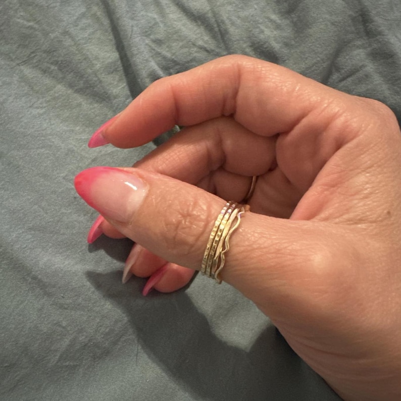 May include: A set of four gold-toned stacking rings with different textures. The rings are worn on the thumb of a hand with pink and white ombre nail polish.