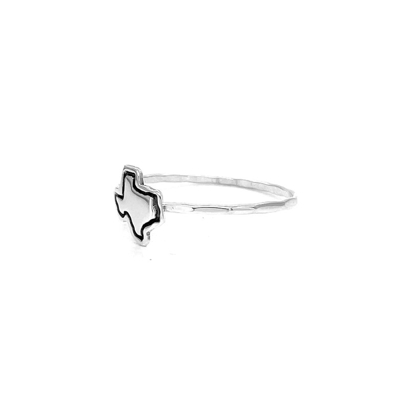 May include: A silver ring featuring the outline of the state of Texas. The ring has a hammered band and a raised Texas shape with a black border. The ring is shown on a white background.