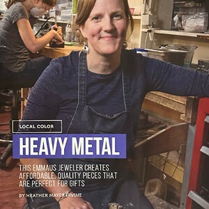 May include: A woman wearing a blue denim overall dress and a blue shirt is working in a jewellery studio. She is holding a hammer in her hand. The text "LOCAL COLOR HEAVY METAL" is displayed above her. The text "THIS EMMAUS JEWELLER CREATES AFFORDABLE, QUALITY PIECES THAT ARE PERFECT FOR GIFTS BY HEATHER MAYER IRVINE" is displayed below her.