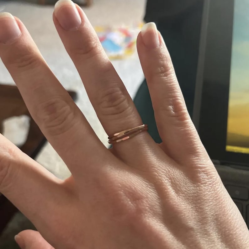 K&ouml;nnte beinhalten: Ein ros&eacute;goldener Ring an einem Finger. Der Ring hat ein minimalistisches Design mit zwei d&uuml;nnen B&auml;ndern. Die B&auml;nder sind durch kleine, rechteckige Akzente verbunden. Der Ring ist ein schlichtes, aber elegantes Schmuckst&uuml;ck.