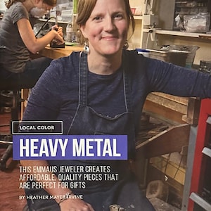 K&ouml;nnte beinhalten: Eine Frau in einer blauen Jeanssch&uuml;rze l&auml;chelt und h&auml;lt einen Hammer in einer Werkstatt. Der Text "HEAVY METAL" wird in einem blauen Feld angezeigt, mit dem Text "THIS EMMAUS JEWELER CREATES AFFORDABLE, QUALITY PIECES THAT ARE PERFECT FOR GIFTS".