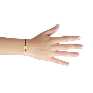 May include: A simple bracelet with a small, round, gold charm on a dark red cord.