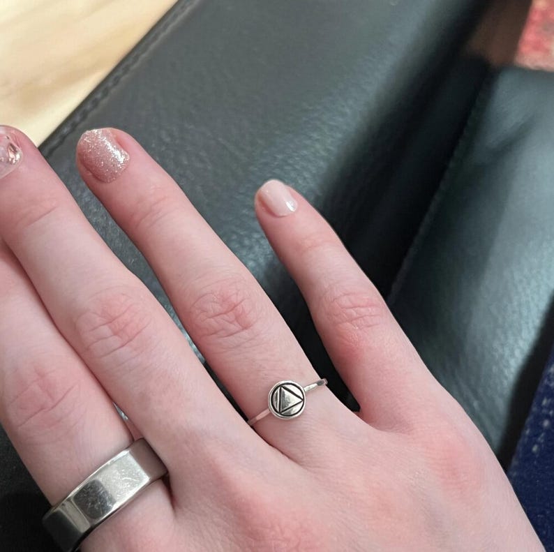 May include: A silver ring with a small, round, silver charm featuring a triangle design. The ring is on a person's finger.