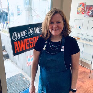 May include: A woman wearing a black t-shirt and a blue denim overall dress with a variety of colorful buttons. She is wearing a layered necklace with beads and stones. The sign behind her says "Come in We're Awesome".