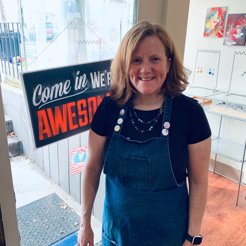 May include: A woman wearing a black t-shirt and a blue denim overall dress with a variety of colorful buttons. She is wearing a layered necklace with beads and stones. The sign behind her says "Come in We're Awesome".