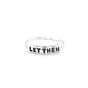 Può includere: Un anello d'argento con le parole "LET THEM" incise su una fascia.