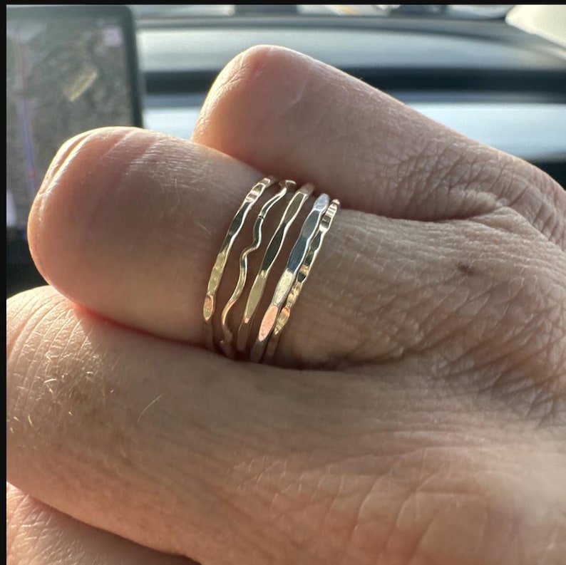 May include: A set of five gold-toned stacking rings. The rings are all different widths and have a hammered texture. The rings are worn on a finger.