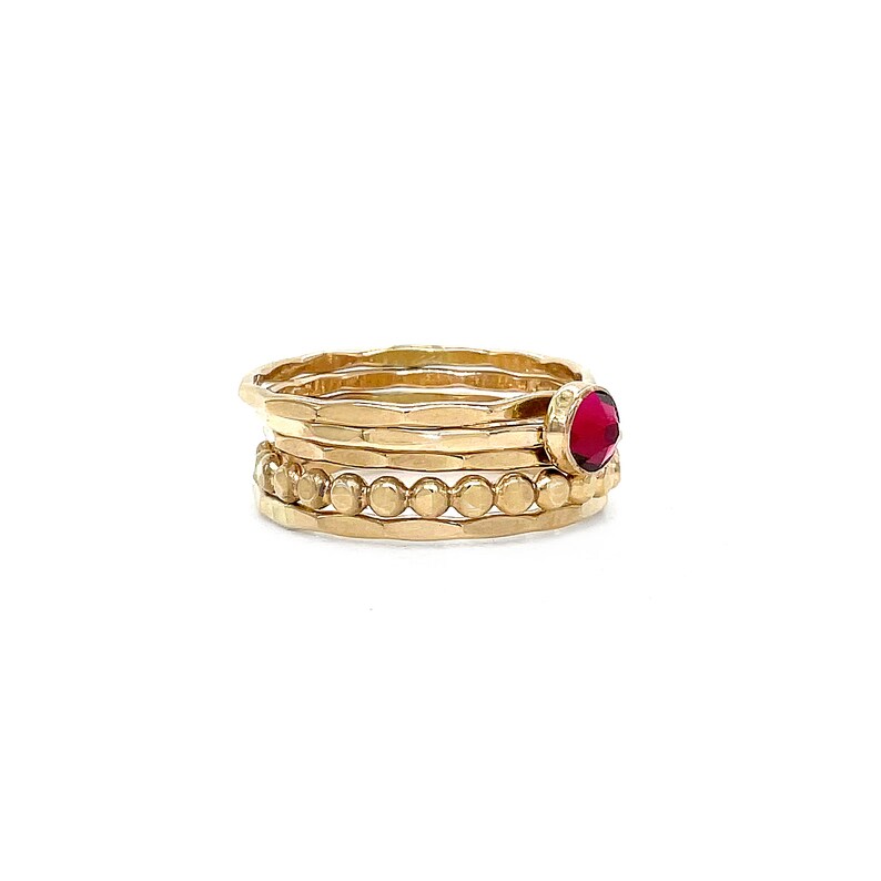 May include: A set of four gold rings, one with a red gemstone, stacked on a white background.