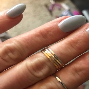 May include: A hand wearing a stack of five thin gold rings on the ring finger. The hand is also wearing a silver ring on the pinky finger.