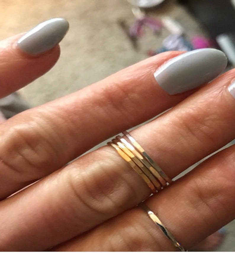 May include: A hand wearing a stack of five thin gold rings on the ring finger. The hand is also wearing a silver ring on the pinky finger.