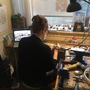 May include: A person wearing a face mask is working on a piece of jewelry with a grinder. The person is sitting at a workbench with various tools and materials. There is a laptop computer on the workbench with a screen showing a grid of images.