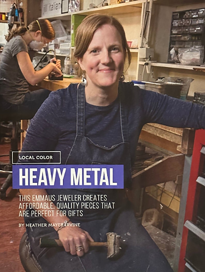 May include: A woman wearing a blue denim overall dress and a blue shirt is working in a jewelry studio. She is holding a hammer in her hand. The text "LOCAL COLOR HEAVY METAL" is displayed above her. The text "THIS EMMAUS JEWELER CREATES AFFORDABLE, QUALITY PIECES THAT ARE PERFECT FOR GIFTS BY HEATHER MAYER IRVINE" is displayed below her.