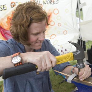 May include: A person uses a hammer to shape metal. The hammer has a yellow handle and a black head. The person is wearing a blue shirt and a watch. The background is a white tent with the words "House of Metal Works" printed on it.