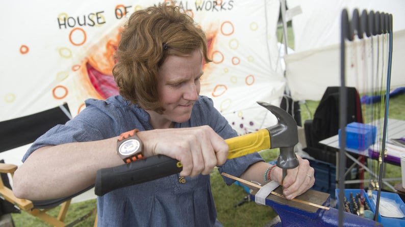 May include: A person uses a hammer to shape metal. The hammer has a yellow handle and a black head. The person is wearing a blue shirt and a watch. The background is a white tent with the words "House of Metal Works" printed on it.