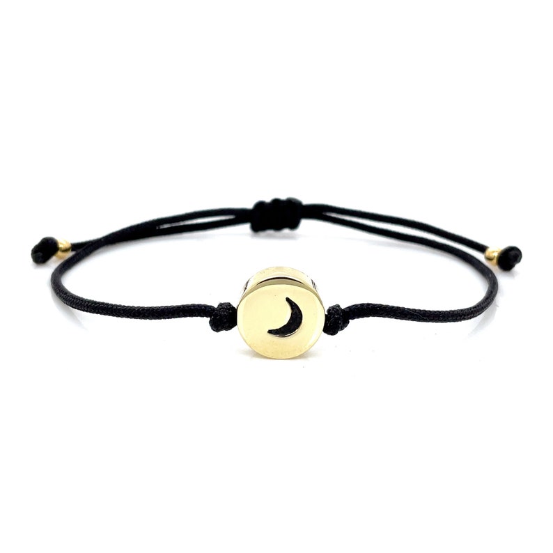 May include: A black string bracelet with a gold circular charm featuring a crescent moon design.