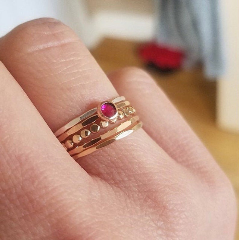 May include: A set of four gold rings with a single red gemstone in the center. The rings are stacked on top of each other and have a hammered texture.