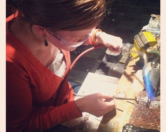 Puede incluir: Una mujer con gafas de seguridad y una camisa roja está trabajando en un proyecto de elaboración de joyas. Está usando un soplete para fundir metal y crear una pieza de joyería.