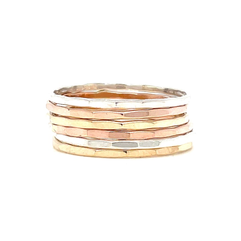 May include: Stack of hammered metal rings in silver, rose gold, and yellow gold. The rings have a textured surface and are stacked together. The rings are displayed on a white background.