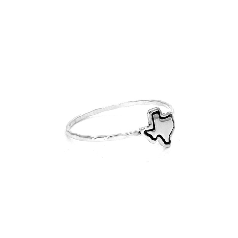 May include: A silver ring featuring a Texas-shaped charm with a black outline. The ring has a hammered texture on the band and is set against a white background. The charm is a detailed representation of the state's outline.